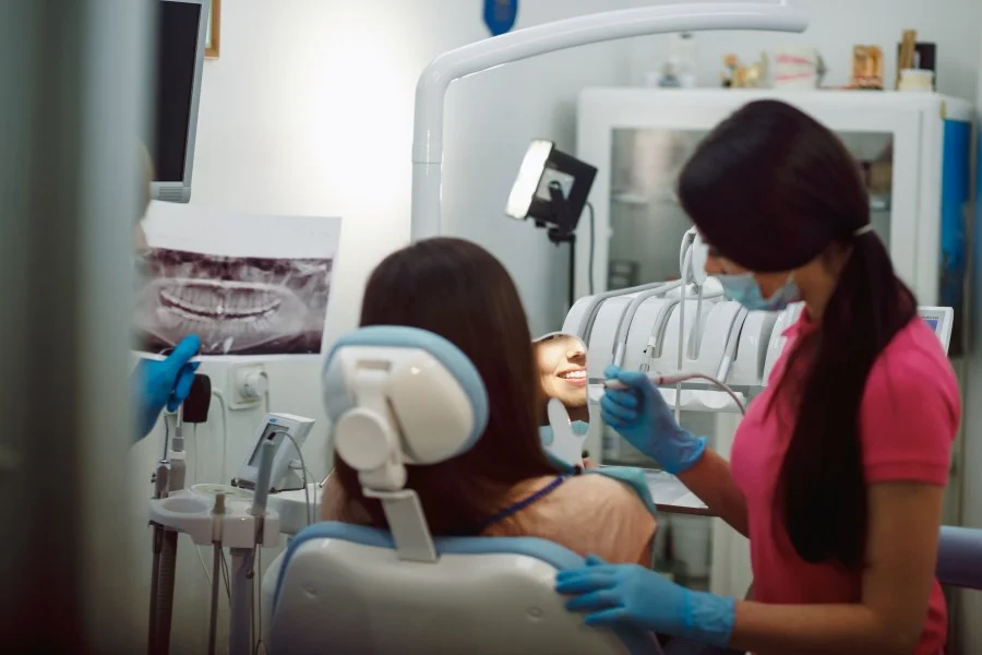 Dentist showing a patient seated in a dental chair while a masked dentist performs an examination. A dental assistant in pink scrubs examining a patient in a dental clinic while an assistant prepares instruments