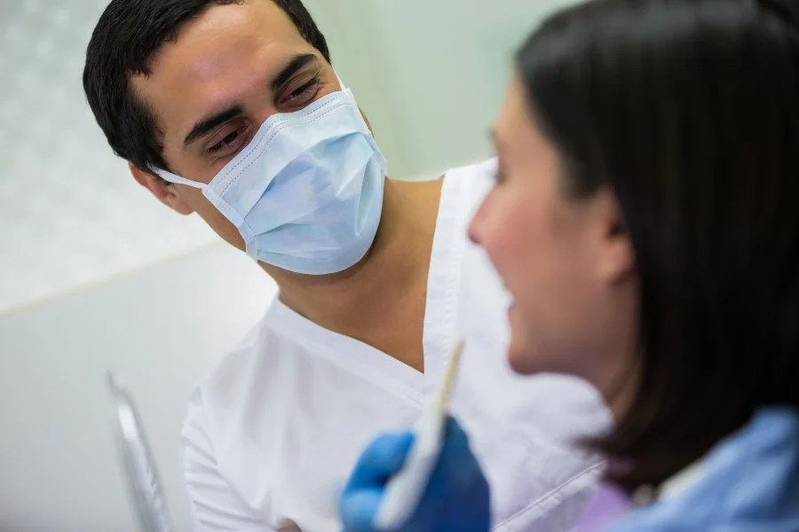 Dentist in a surgical mask examining a female patient during a dental appointment.