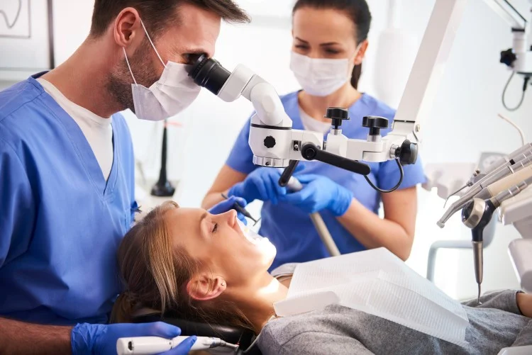 Alt text: Dentist examining a patient’s teeth with a dental microscope while an assistant provides support
